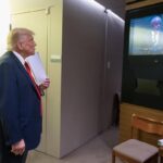 A imagem mostra um homem com cabelo loiro e terno escuro, que parece ser Donald Trump, assistindo a uma apresentação em uma tela. Ele está em um ambiente interno, com uma parede clara ao fundo e uma mesa com papéis e uma planta ao lado. Um homem de terno escuro está atrás dele, observando. Na tela, um orador está falando, mas não é possível identificar quem é.