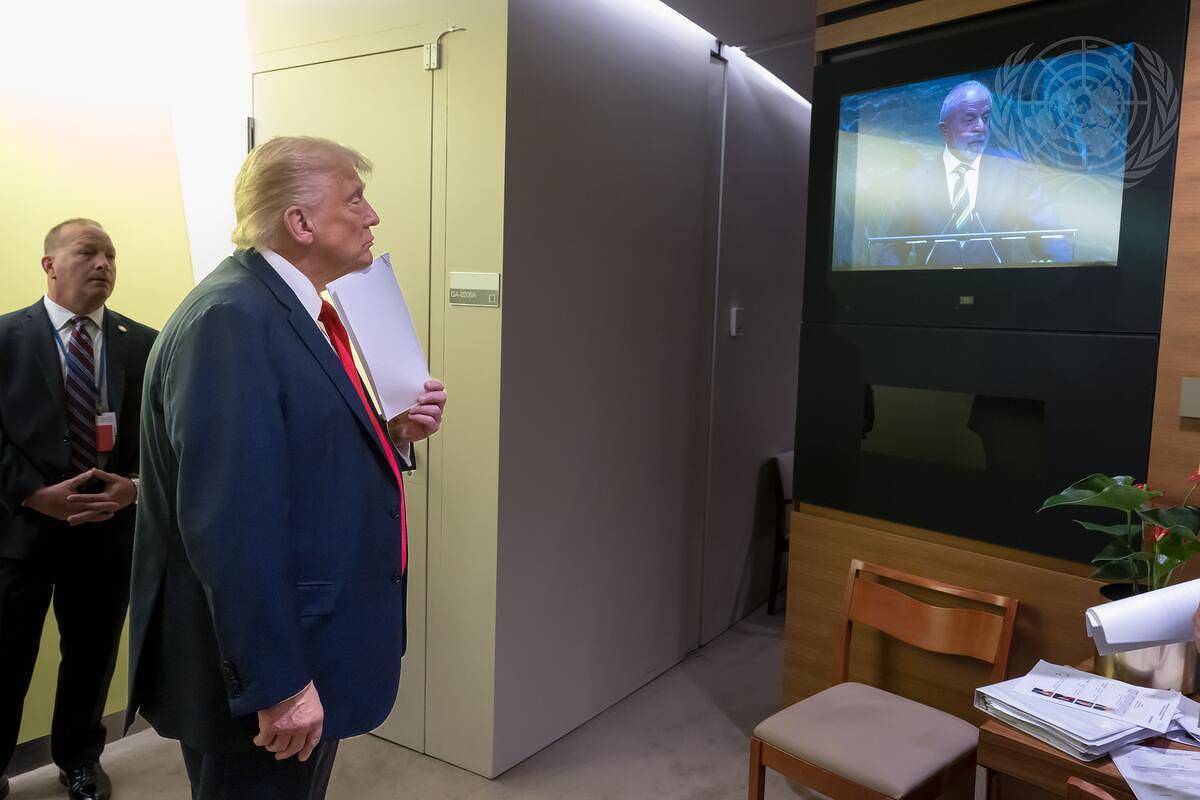 A imagem mostra um homem com cabelo loiro e terno escuro, que parece ser Donald Trump, assistindo a uma apresentação em uma tela. Ele está em um ambiente interno, com uma parede clara ao fundo e uma mesa com papéis e uma planta ao lado. Um homem de terno escuro está atrás dele, observando. Na tela, um orador está falando, mas não é possível identificar quem é.
