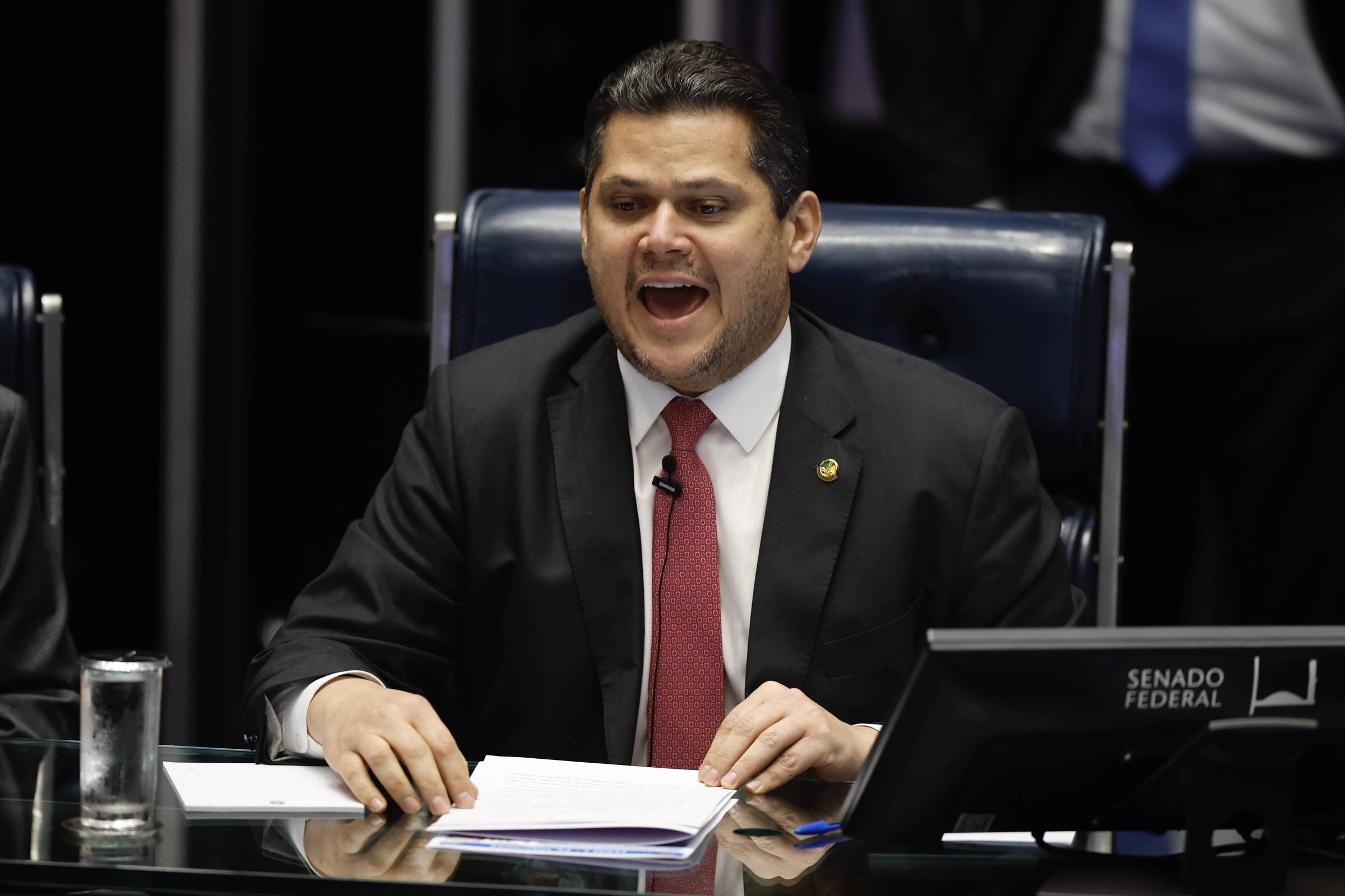 Senador em terno preto e gravata vermelha fala com expressão aberta sentado em cadeira azul no plenário do Senado Federal. À sua frente, mesa com documentos, copo de água e microfone.