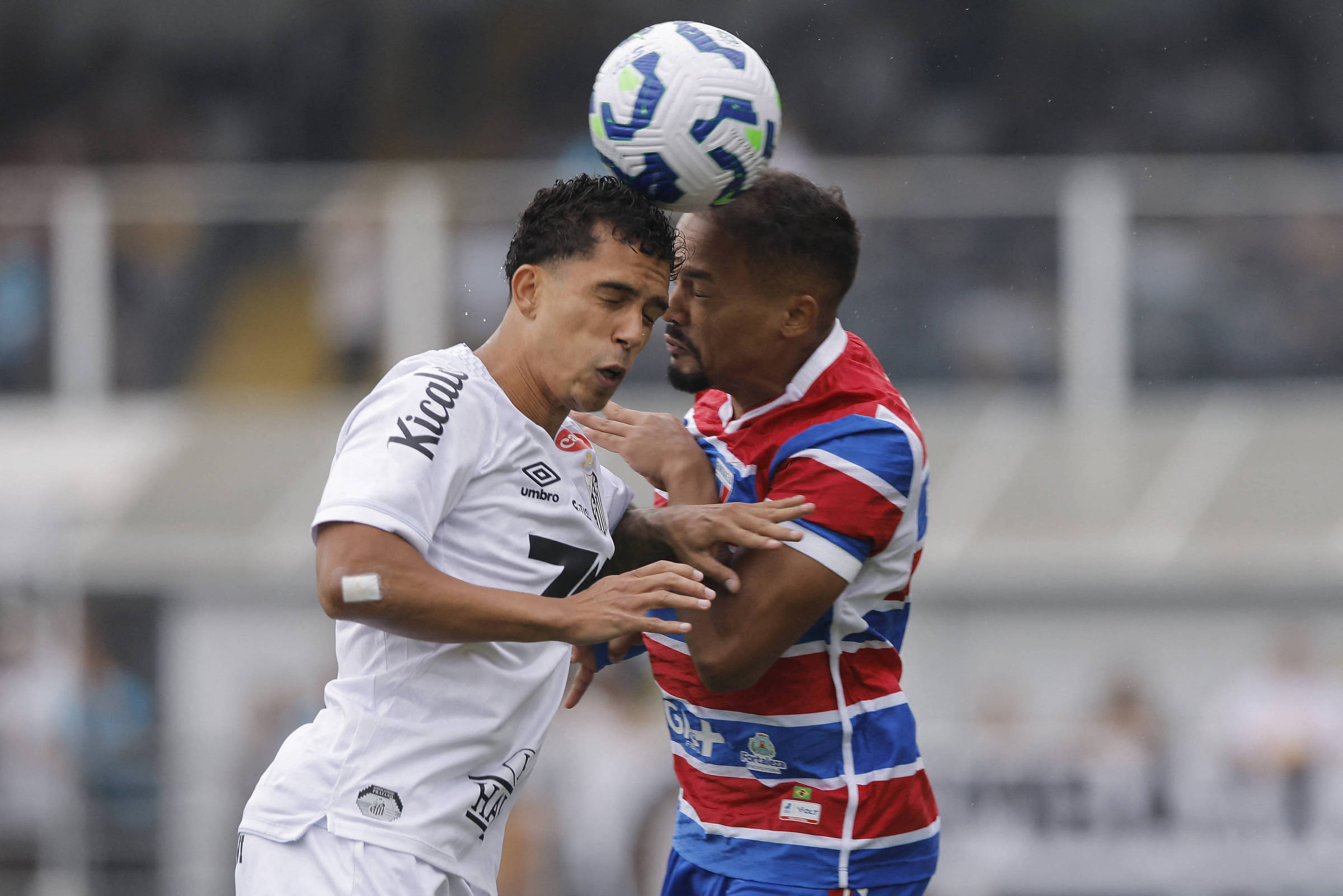 Dois jogadores de times adversários disputam a bola com a cabeça durante jogo de futebol. Um veste uniforme branco e o outro, uniforme listrado em vermelho, azul e branco. O fundo mostra arquibancada desfocada.