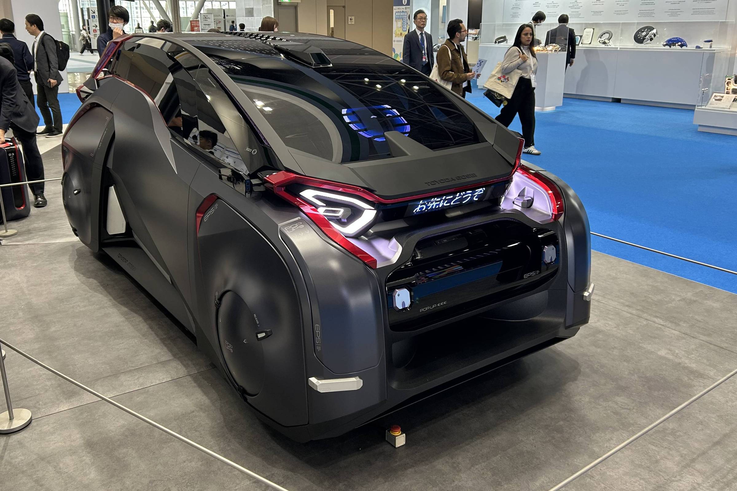 Veículo conceito preto fosco com design aerodinâmico e rodas cobertas exposto em estande de feira. Pessoas circulam ao fundo em ambiente interno com piso cinza e azul.