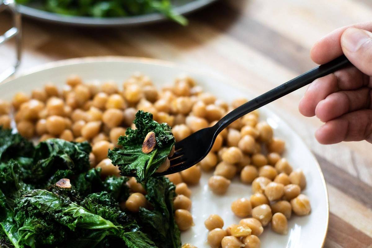A imagem mostra um prato com vegetais verdes, possivelmente couve, e grão-de-bico. Uma mão segura um garfo preto, prestes a pegar um pedaço dos vegetais. O fundo apresenta uma mesa de madeira e um prato adicional com folhas verdes.