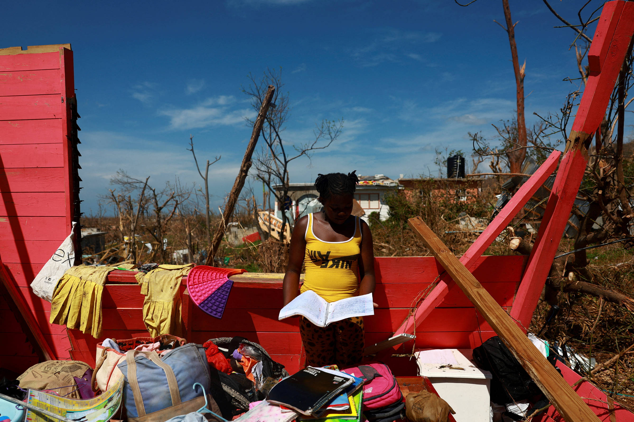 Menina negra vestindo camiseta regata amarela segura caderno em meio a destroços de uma casa vermelha destruída. Ao redor, roupas, mochilas e objetos pessoais espalhados. Céu azul e árvores secas ao fundo.