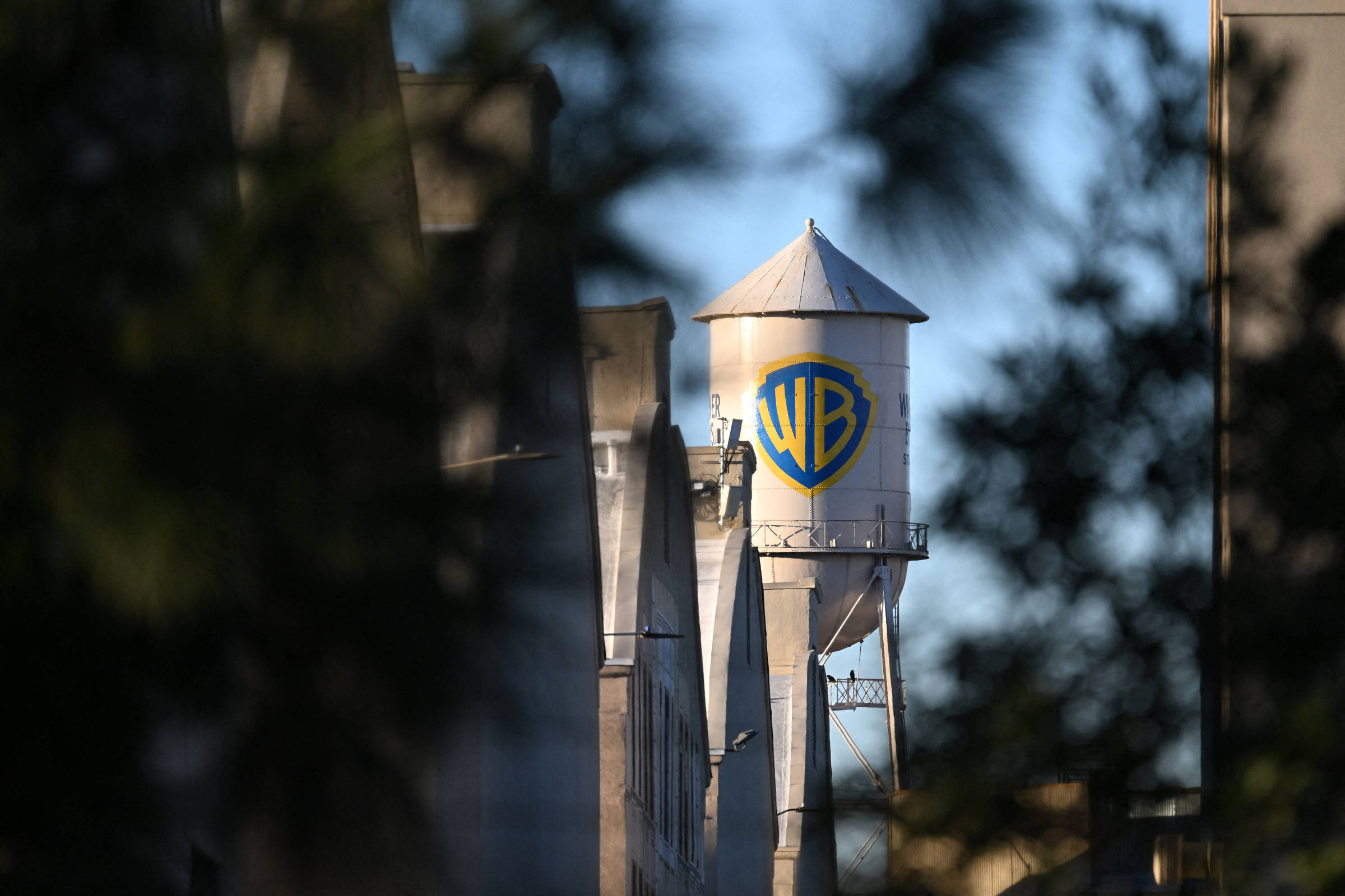 Torre de água branca com o logo azul e amarelo da Warner Bros visível entre prédios, com céu azul ao fundo e vegetação desfocada nas bordas.