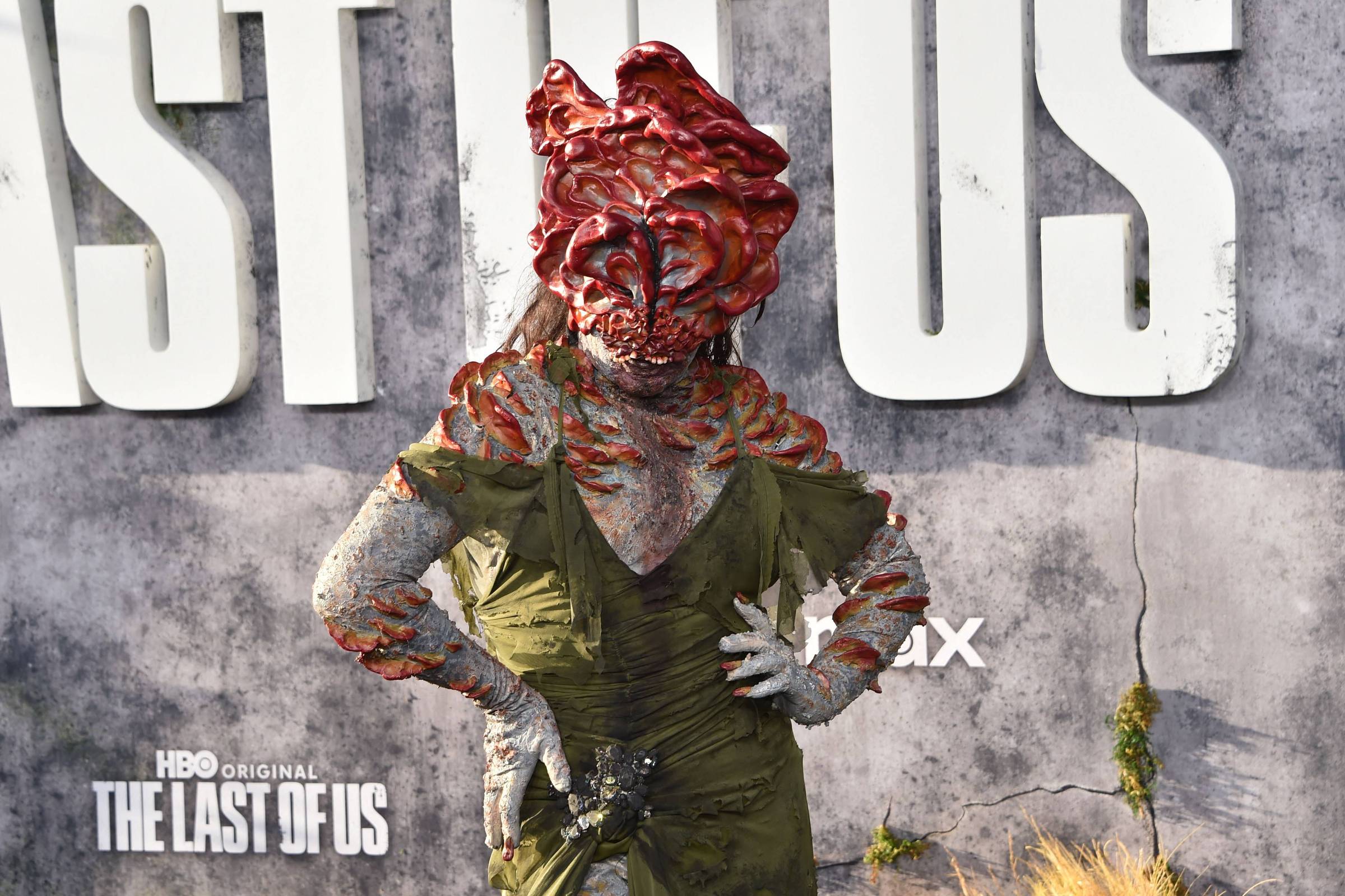 Pessoa vestida como personagem infectado com máscara e maquiagem detalhada simulando fungos, usando vestido verde, posando com mão na cintura em frente a painel com logo de The Last of Us.