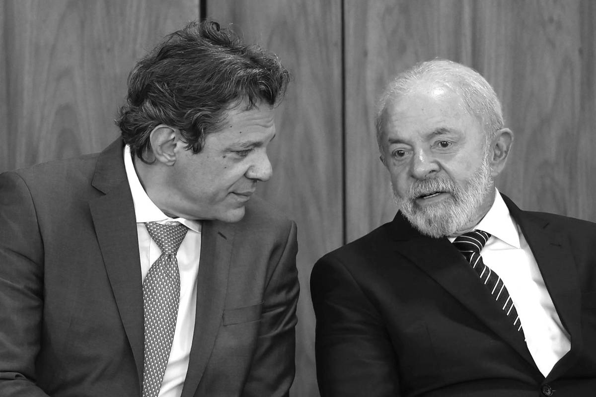 Dois homens vestidos com ternos escuros sentam lado a lado em cadeiras diante de painel de madeira. O homem à esquerda usa gravata vermelha e parece falar com o homem à direita, que usa gravata listrada e escuta atentamente com as mãos entrelaçadas.