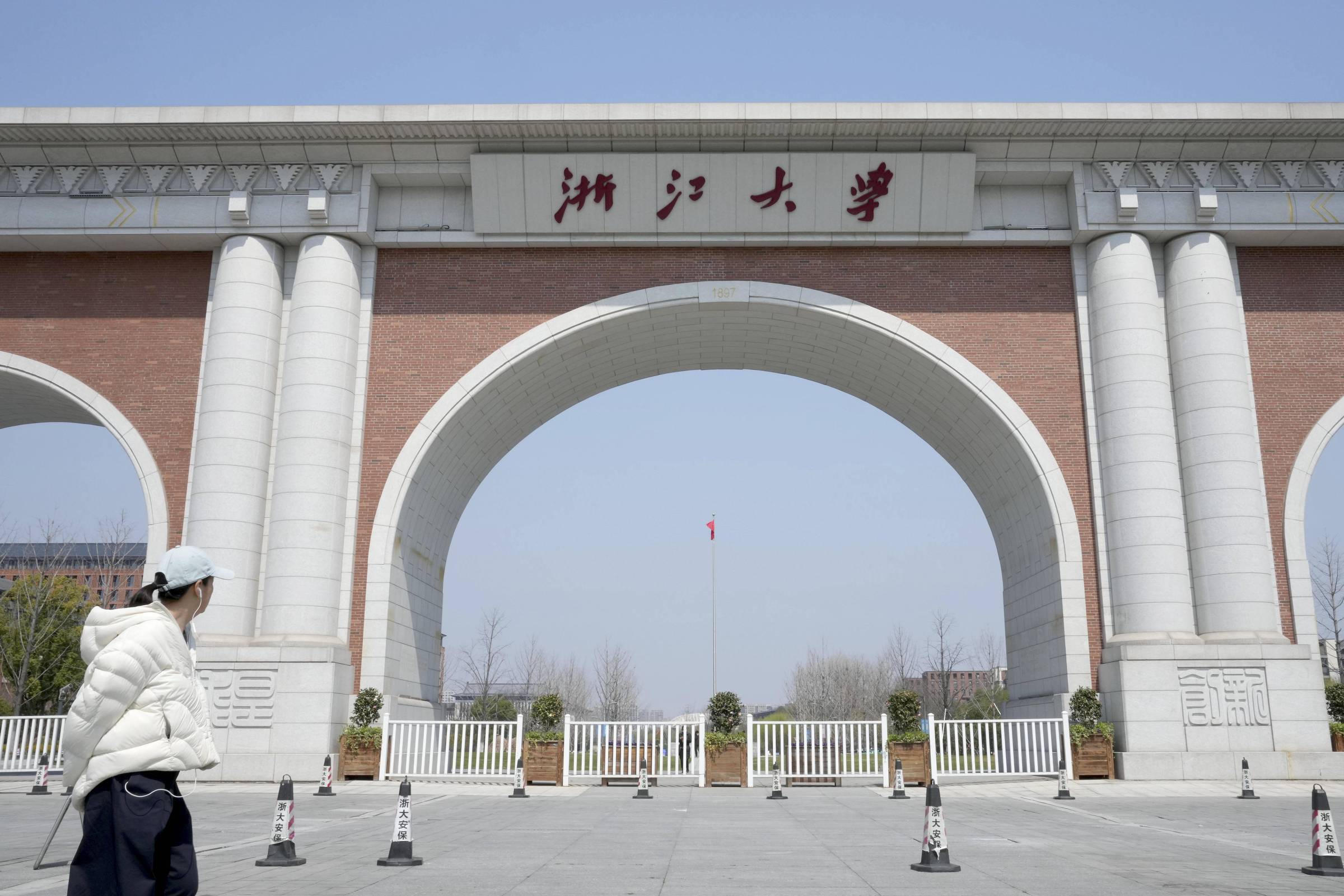 Arco monumental de tijolos vermelhos com colunas brancas na entrada da Universidade de Zhejiang, sob céu azul claro. Pessoa com jaqueta branca e boné caminha à esquerda.