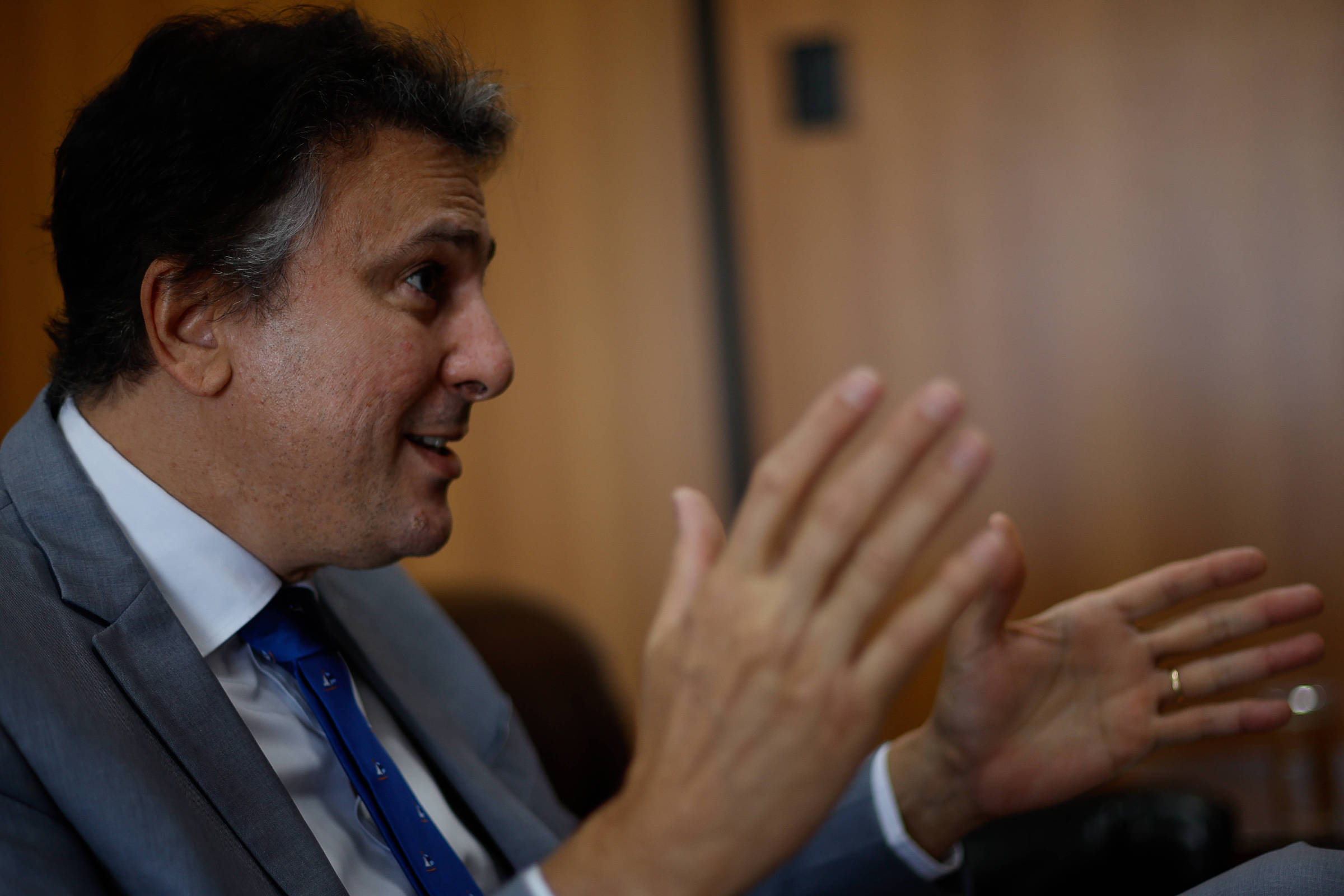 Homem de perfil esquerdo veste terno cinza, camisa branca e gravata azul, gesticulando com as mãos abertas em ambiente interno com parede de madeira ao fundo.