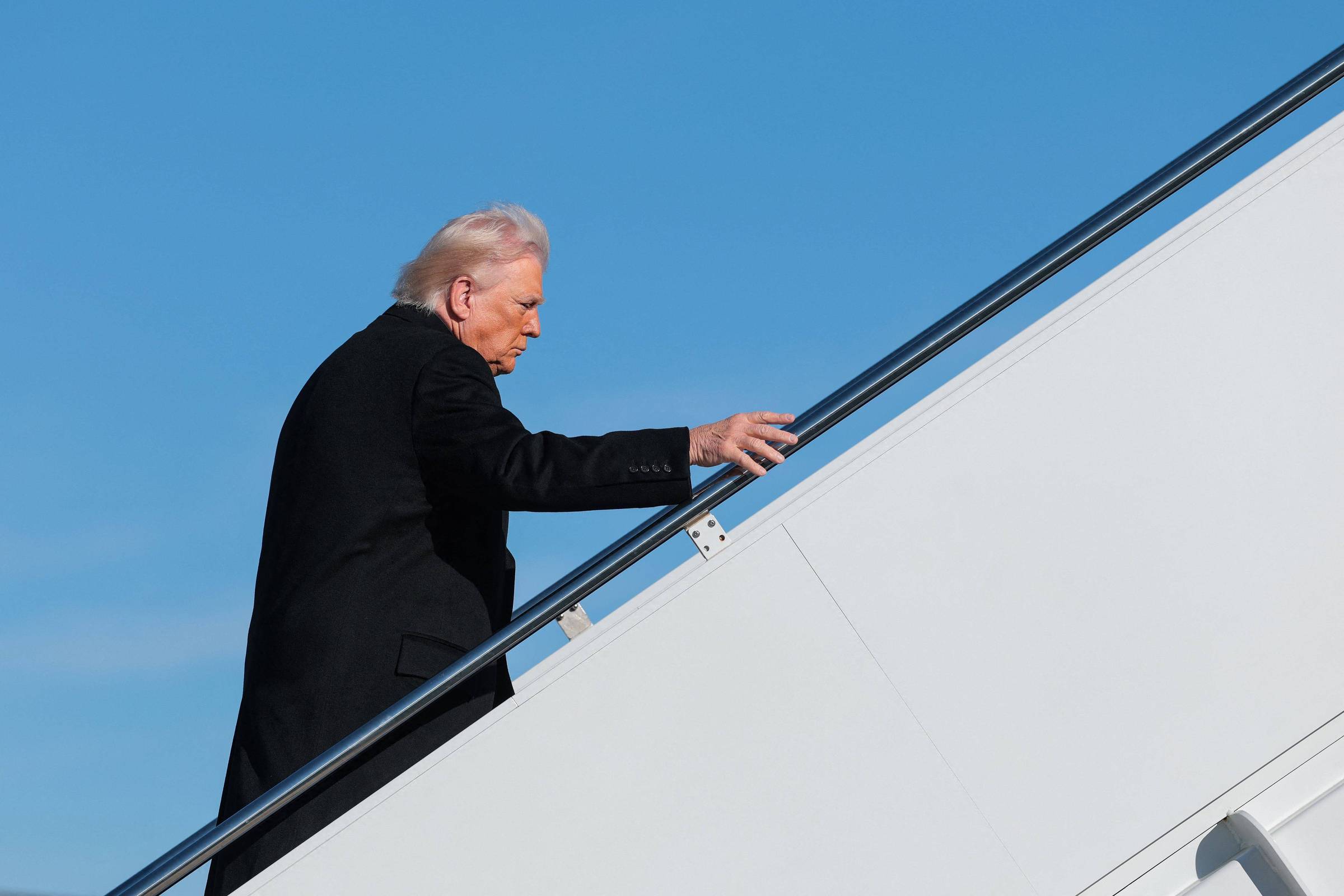 Homem de cabelos grisalhos e sobretudo preto sobe escada de embarque de avião contra céu azul claro.