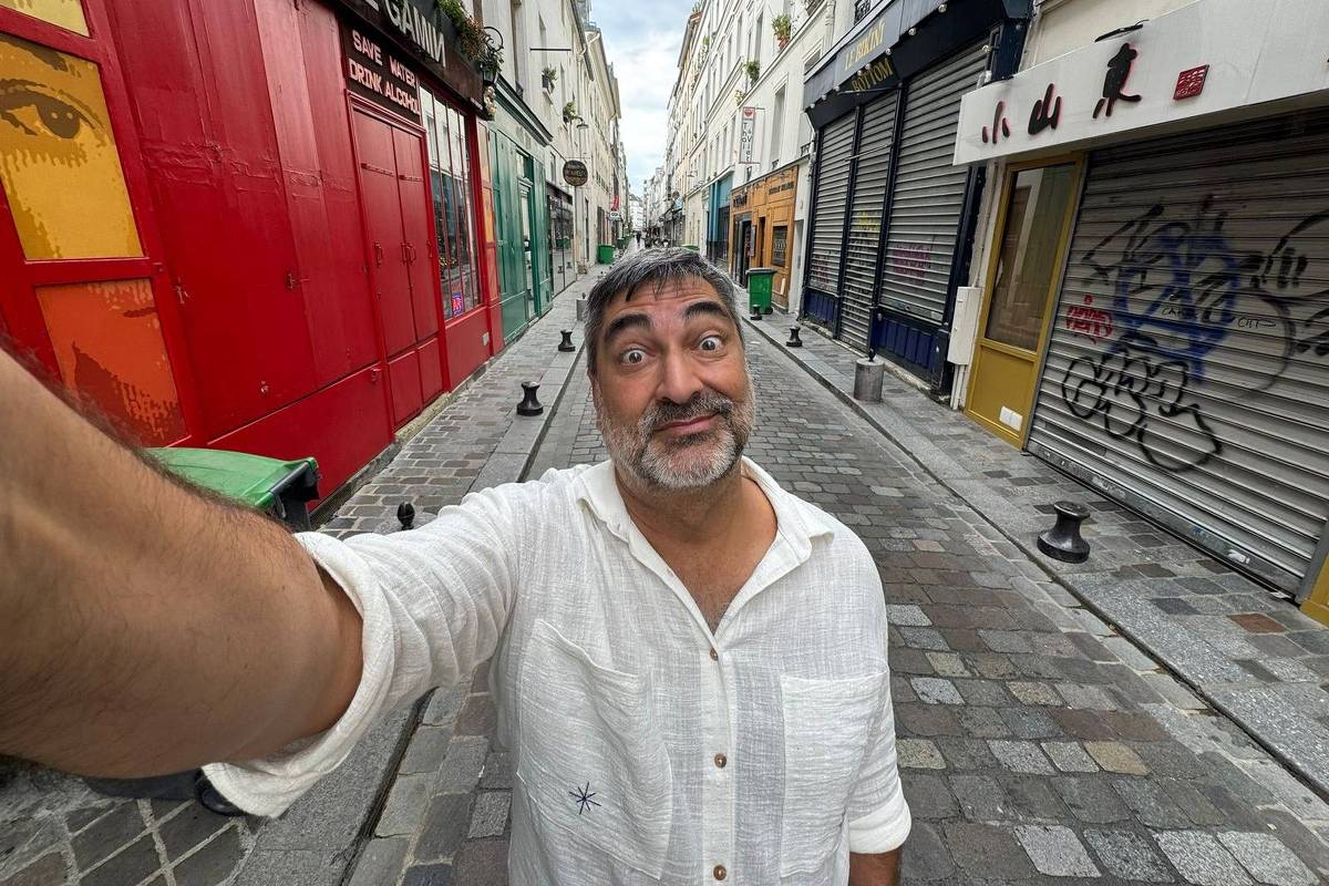 Um homem está tirando uma selfie em uma rua estreita de Paris. Ele tem cabelo grisalho e está usando uma camisa branca. Ao fundo, há prédios com fachadas coloridas, incluindo uma parede vermelha e lojas com portas fechadas. O céu está nublado e a rua é pavimentada com pedras.