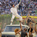 Artista vestido de branco está em pé sobre o capô de um veículo antigo, apontando para cima com a mão direita. Ao redor, grupo diversificado de pessoas com roupas em tons terrosos dança e levanta os braços. Ao fundo, multidão grande em arquibancada e vegetação alta