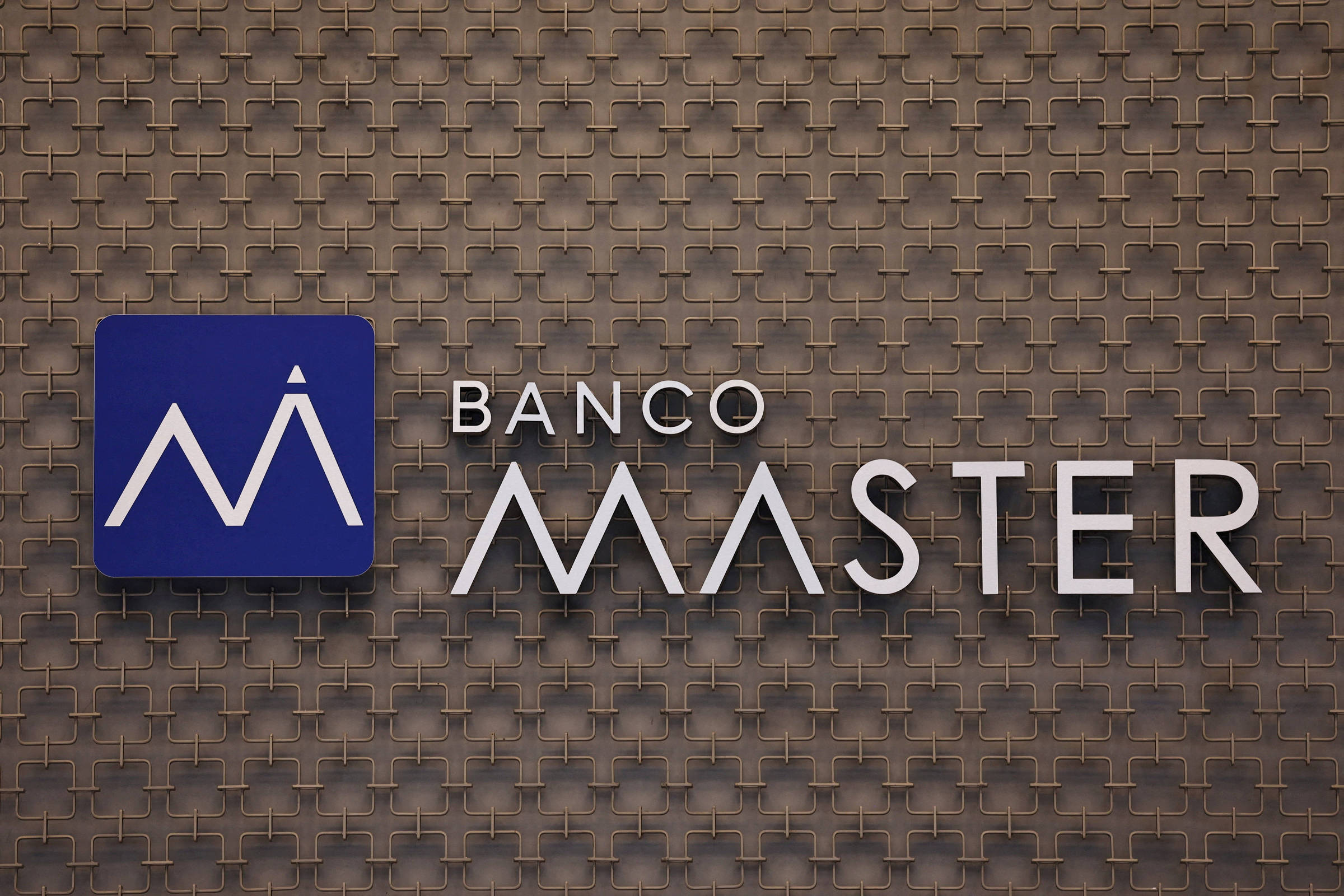 Logotipo do Banco Master fixado em parede com textura quadriculada marrom. À esquerda, símbolo branco estilizado em fundo azul; à direita, nome
