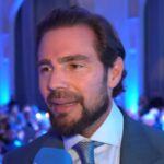 Homem de barba e cabelo escuro veste terno azul e gravata clara, falando ao microfone azul em ambiente interno com iluminação azul e público ao fundo.