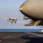 Avião decolando da pista de um porta-aviões sobre o mar, com caça estacionado em primeiro plano à direita. Céu claro e mar calmo ao fundo.