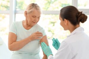 Uma mulher idosa, de pele clara e cabelos loiros curtos, recebe uma aplicação de vacina no braço esquerdo. Ela veste uma camiseta verde-clara e olha calmamente para o braço enquanto a profissional de saúde, vista de costas e de perfil, realiza o procedimento. A profissional utiliza um jaleco branco e luvas descartáveis de cor verde. O ambiente é iluminado e sugere um consultório médico, com uma janela ampla ao fundo que mostra árvores desfocadas pela luz solar.
