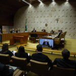 Ministros sentados em semicírculo no plenário do Supremo Tribunal Federal, com público e advogados na plateia. Parede de pedra com cruz e brasão ao fundo.