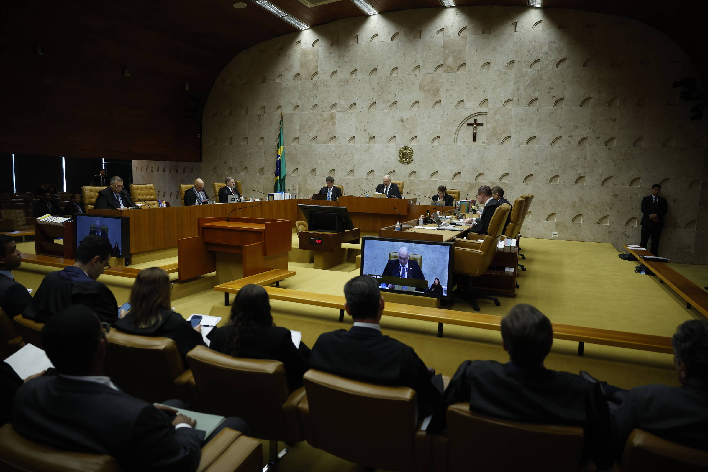 Ministros sentados em semicírculo no plenário do Supremo Tribunal Federal, com público e advogados na plateia. Parede de pedra com cruz e brasão ao fundo.