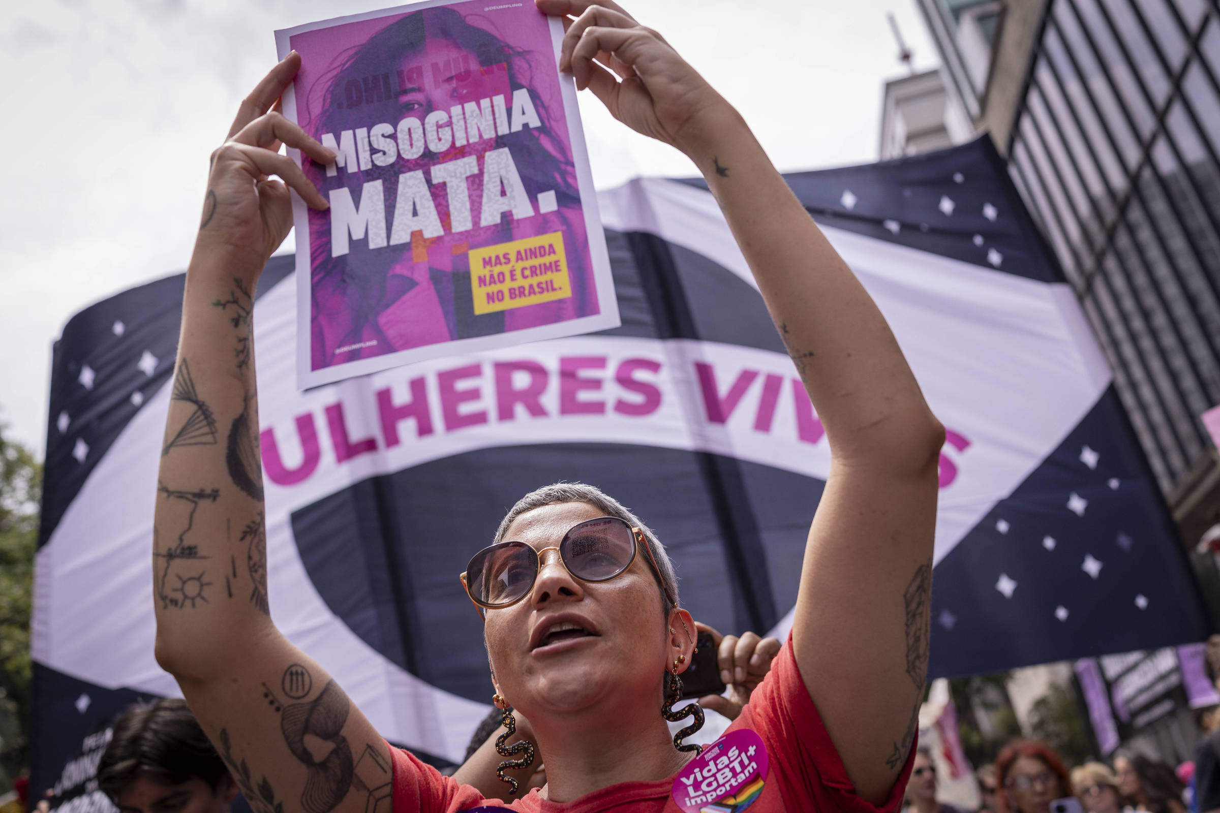 Mulher com óculos escuros e tatuagens levanta cartaz roxo com texto