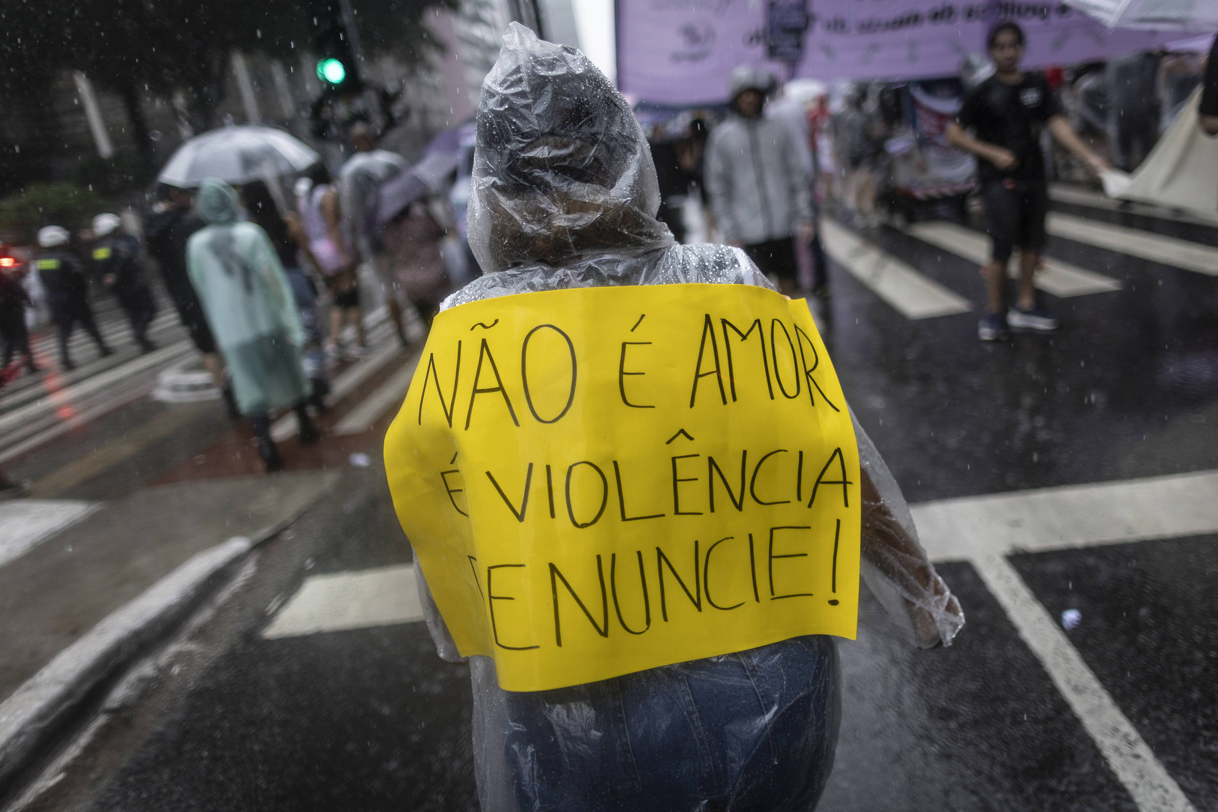 Pessoa vestindo capa de chuva transparente segura cartaz amarelo nas costas com a frase