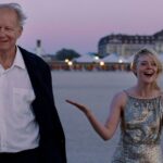 filme-valor-sentimental-casal-caminhando-na-praia-ao-entardecer-com-mulher-em-vestido-brilhante-e-homem-de-blazer