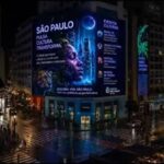 Painel digital iluminado em fachada curva de edifício no centro de São Paulo exibe campanha cultural com rosto feminino e texto sobre eventos culturais. Ruas molhadas refletem luzes da cidade e trânsito de veículos e pedestres é visível à noite.