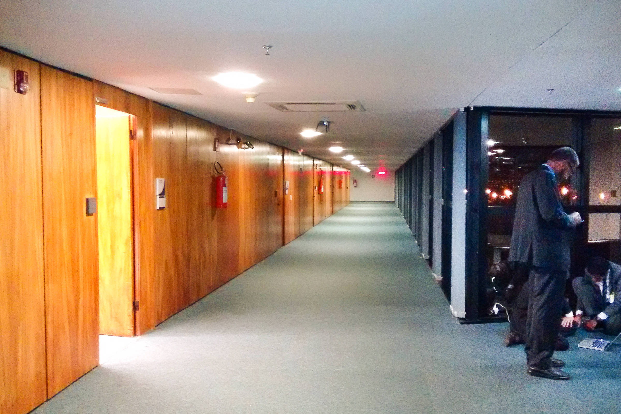 Corredor longo com paredes de madeira à esquerda e janelas grandes à direita. Duas pessoas vestindo ternos estão no canto direito: uma está em pé olhando para um celular e outra está agachada mexendo em um laptop no chão. Ambiente iluminado artificialmente, aparentando ser à noite.