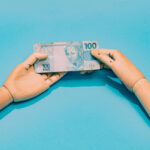 A imagem mostra duas mãos de madeira, uma de cada lado, segurando uma nota de 100 reais. O fundo é azul claro, destacando a nota que possui a figura de uma mulher e o valor impresso em destaque. As mãos estão posicionadas de forma a parecer que estão trocando a nota.