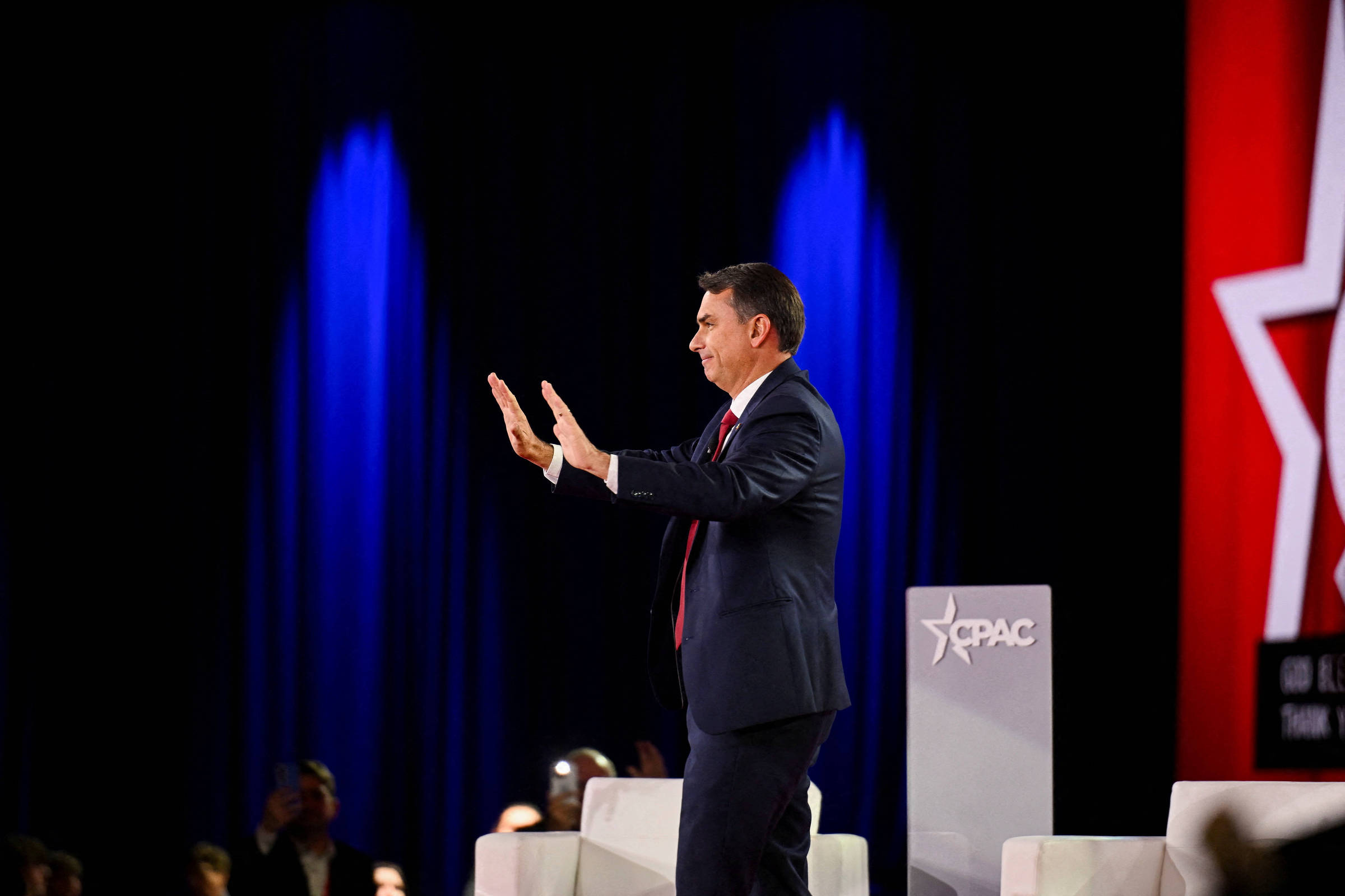Homem de terno escuro e gravata vermelha está de perfil, com as mãos levantadas em gesto de parada, em palco com fundo azul e vermelho iluminado. Ao lado, púlpito com logo do CPAC e cadeiras brancas.