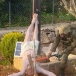 Estátua mostra Jesus pendurado de cabeça para baixo em poste de madeira, com as pernas amarradas ao poste. Soldado com uniforme camuflado e capacete está ao lado, aparentemente manipulando as amarras. Cena ocorre em área externa com vegetação, arbustos e cerca de metal ao fundo.