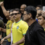 Homem de óculos escuros e camisa amarela da seleção brasileira caminha cercado por várias pessoas, algumas também vestindo amarelo. Um homem ao fundo levanta o braço com o punho fechado. Ambiente externo com vegetação ao fundo.