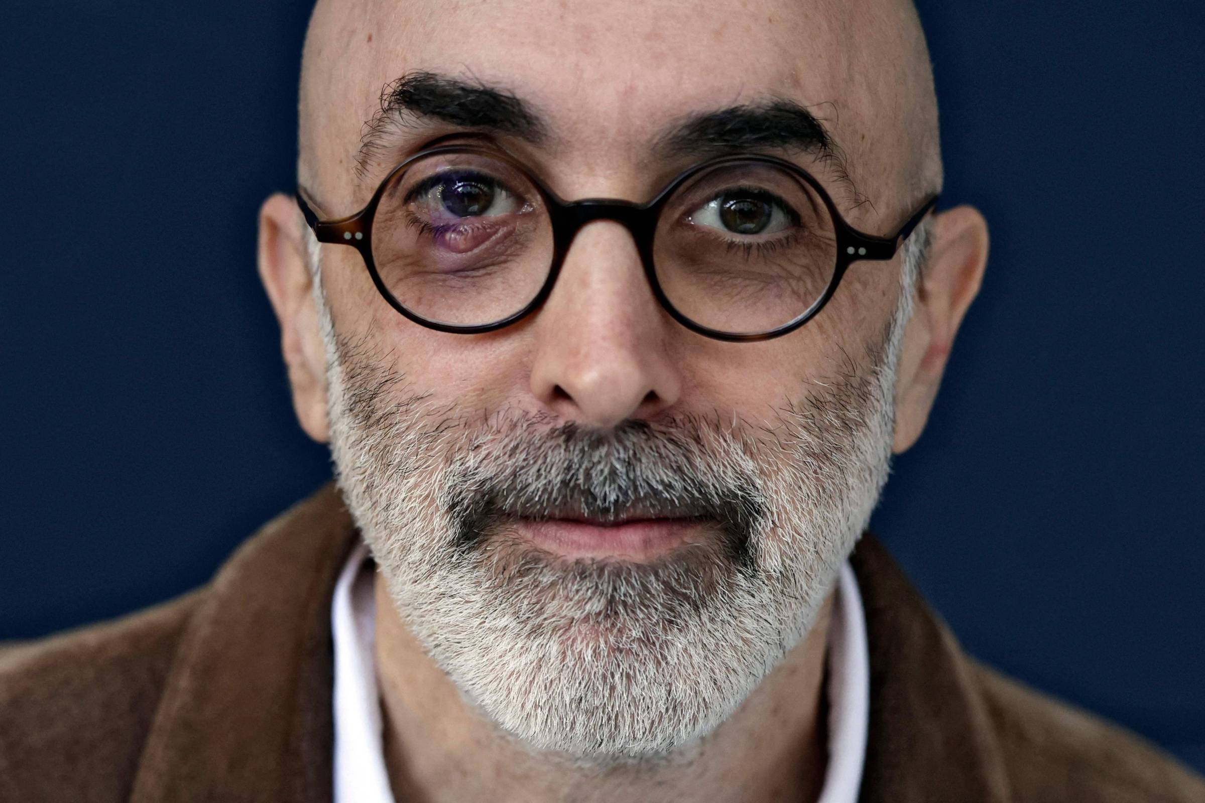 Homem careca com barba grisalha usa óculos redondos de armação preta. Fundo azul escuro e roupa marrom com camisa branca visíveis.