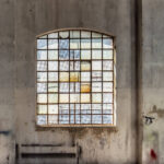 Três janelas grandes com grades de metal e vidros coloridos e quebrados em parede de concreto desgastada. Luz do sol entra pelas janelas, projetando sombras no chão de cimento. Estrutura de madeira do teto visível acima.