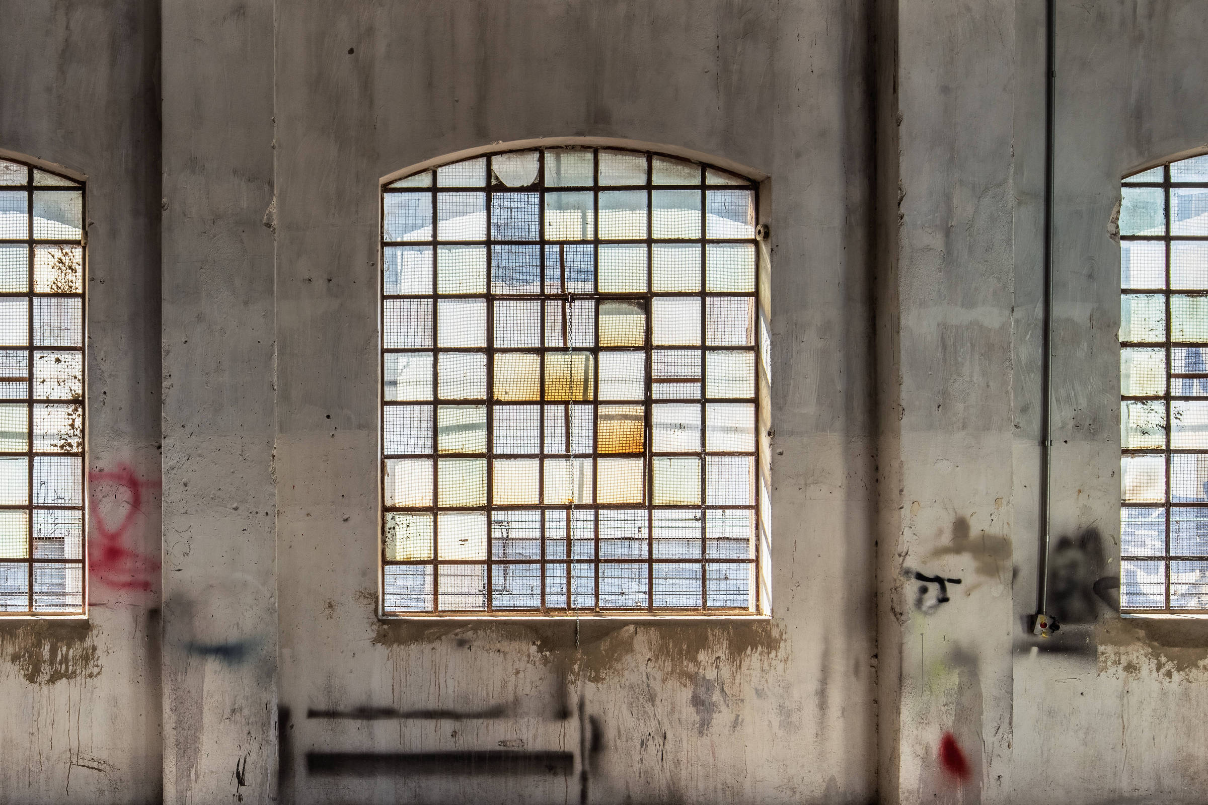 Três janelas grandes com grades de metal e vidros coloridos e quebrados em parede de concreto desgastada. Luz do sol entra pelas janelas, projetando sombras no chão de cimento. Estrutura de madeira do teto visível acima.