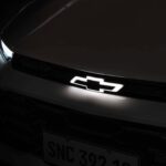 Emblema da Chevrolet iluminado em branco no centro da grade frontal de um carro escuro. Farol esquerdo parcialmente visível com luz acesa. Placa dianteira com caracteres SNC 392 18.