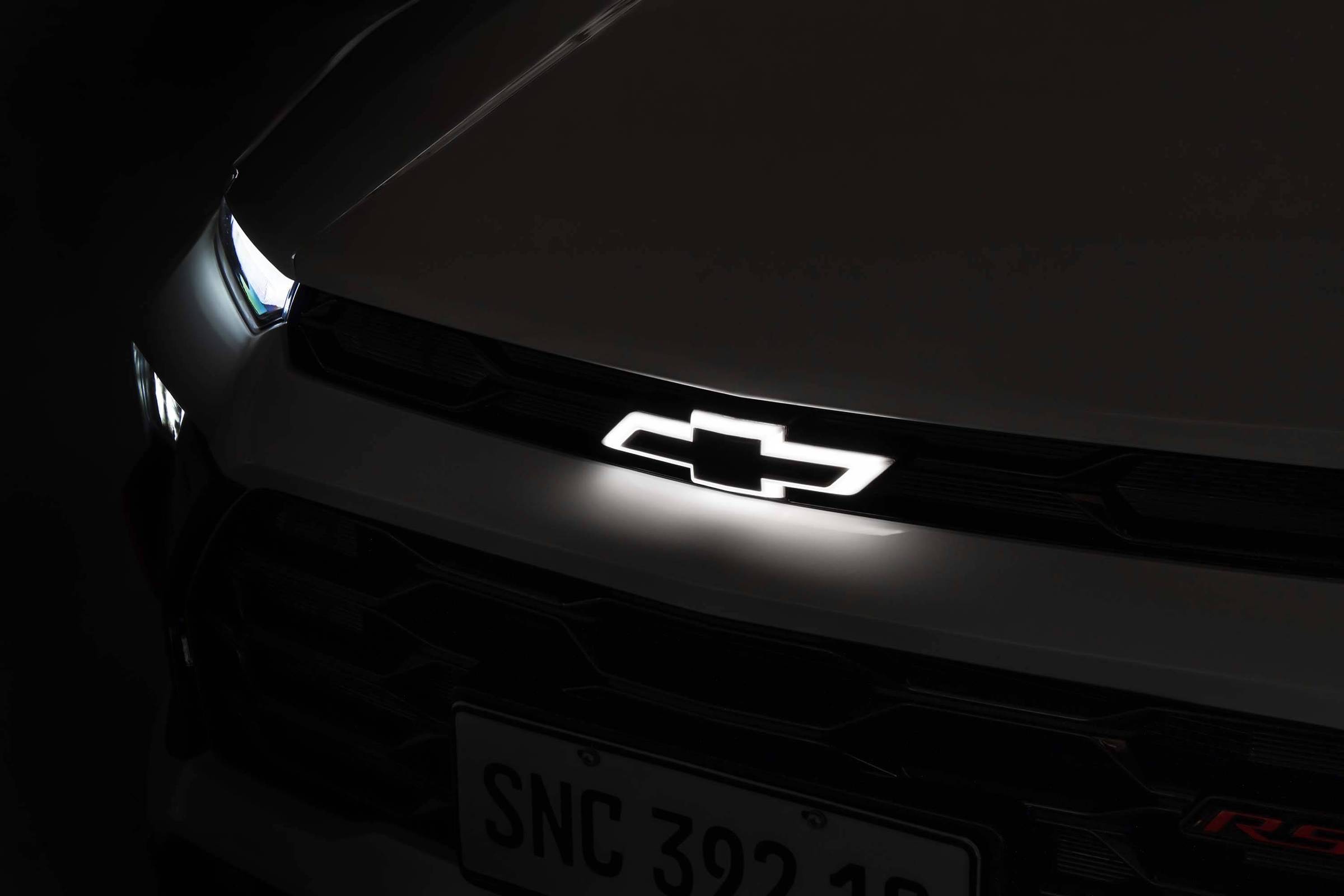 Emblema da Chevrolet iluminado em branco no centro da grade frontal de um carro escuro. Farol esquerdo parcialmente visível com luz acesa. Placa dianteira com caracteres SNC 392 18.