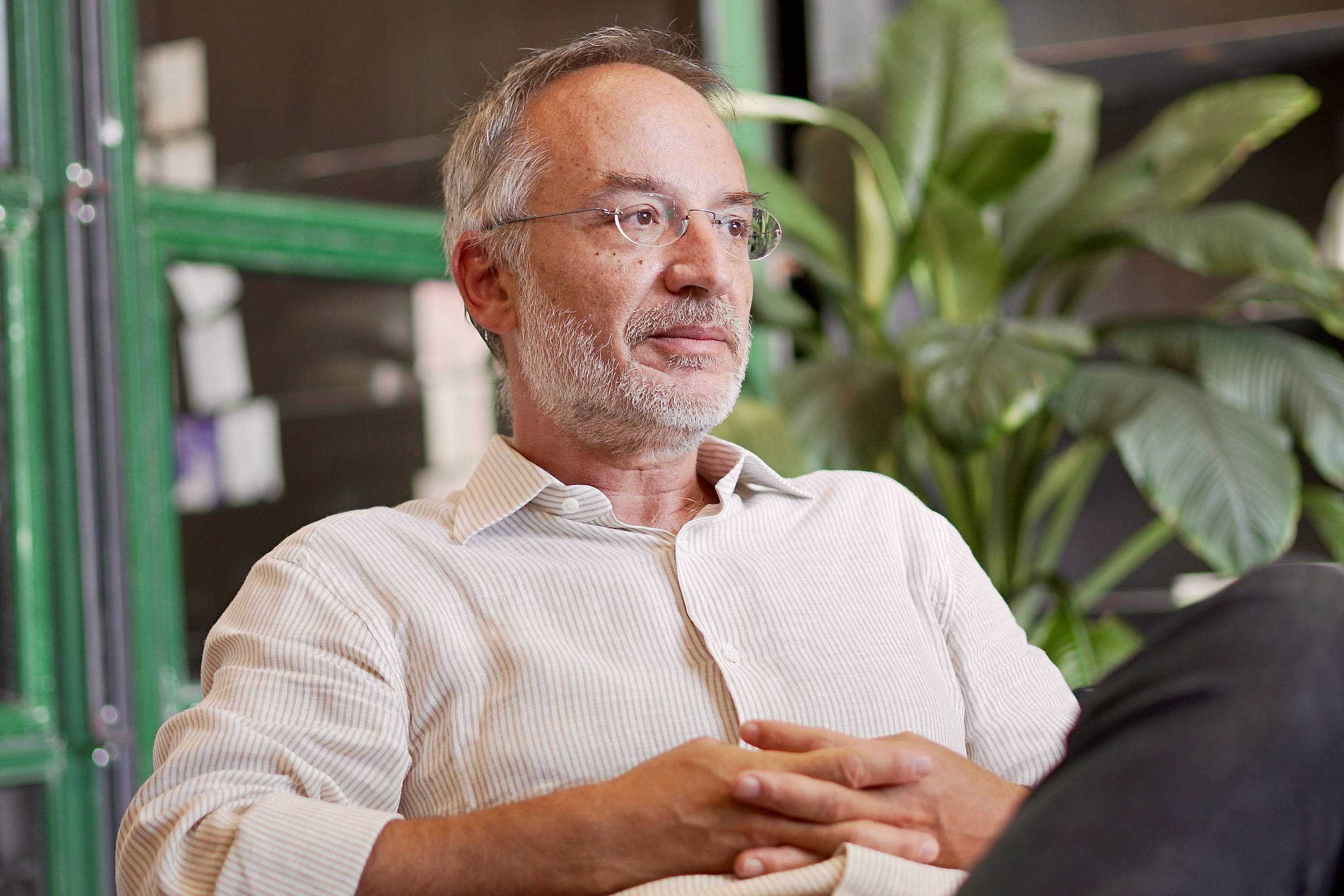 Homem de meia-idade com barba grisalha e óculos, vestido com camisa clara, sentado com as mãos entrelaçadas, olhando para o lado. Ao fundo, plantas verdes e estrutura metálica verde.