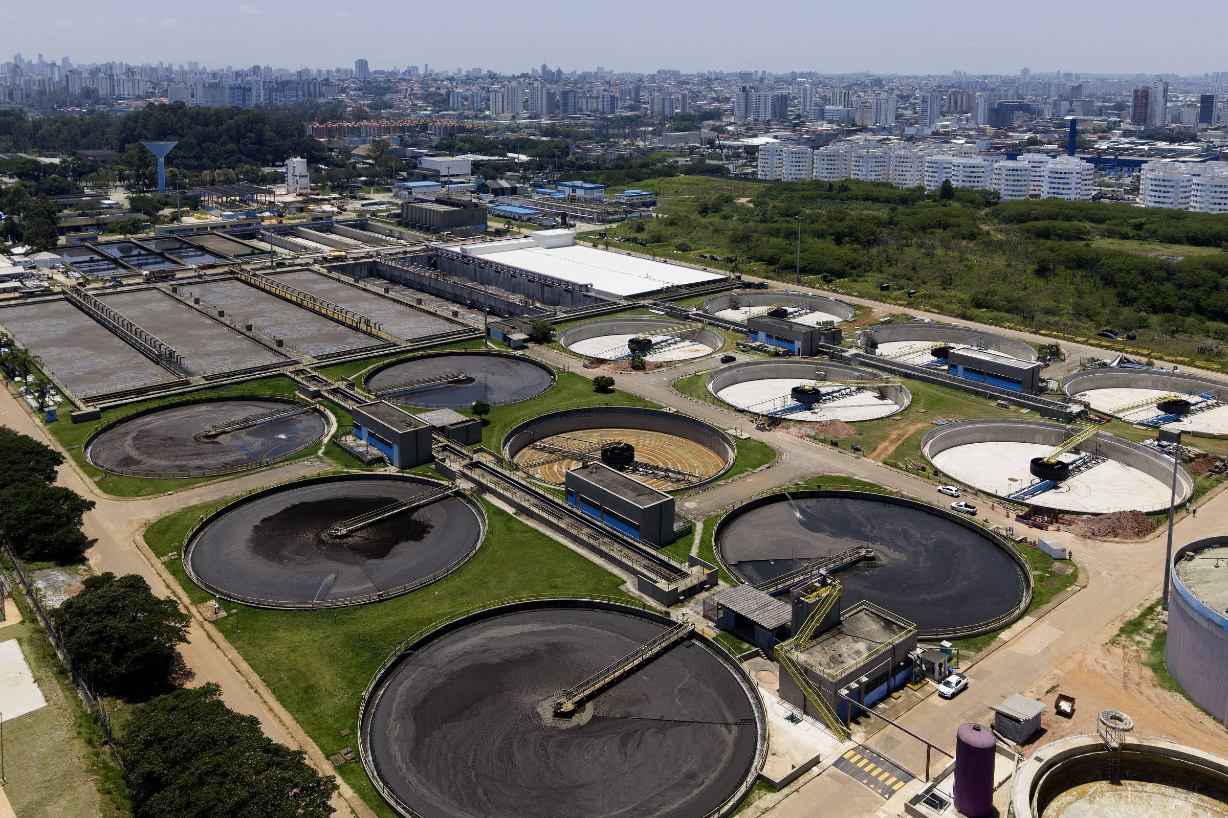 Vista aérea de estação de tratamento de água com vários tanques circulares em diferentes estágios de tratamento. Ao fundo, área urbana com prédios residenciais e vegetação entre a estação e a cidade.