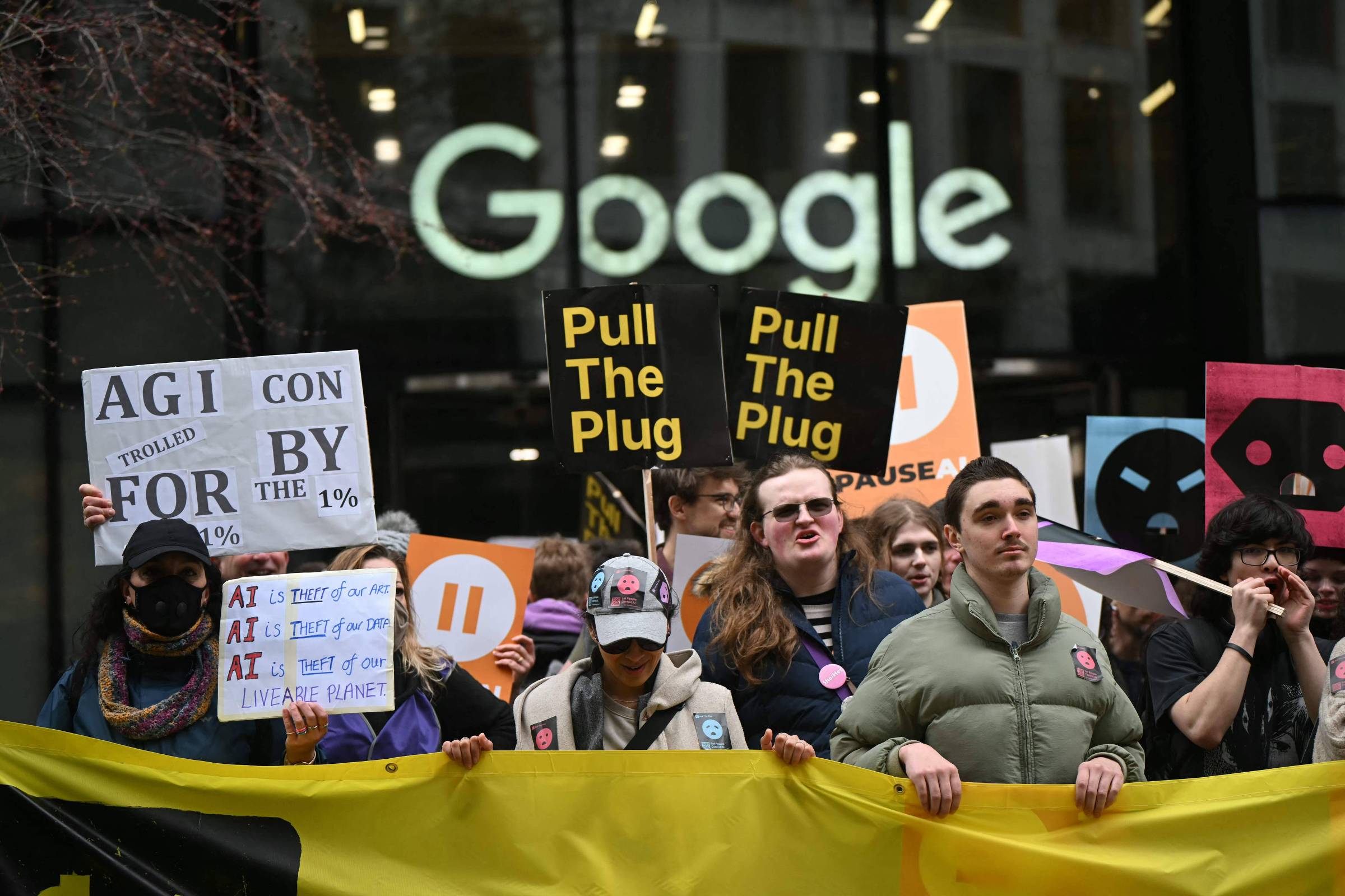 Grupo de manifestantes segura cartazes e faixa amarela em frente à fachada com logo do Google. Placas exibem mensagens como