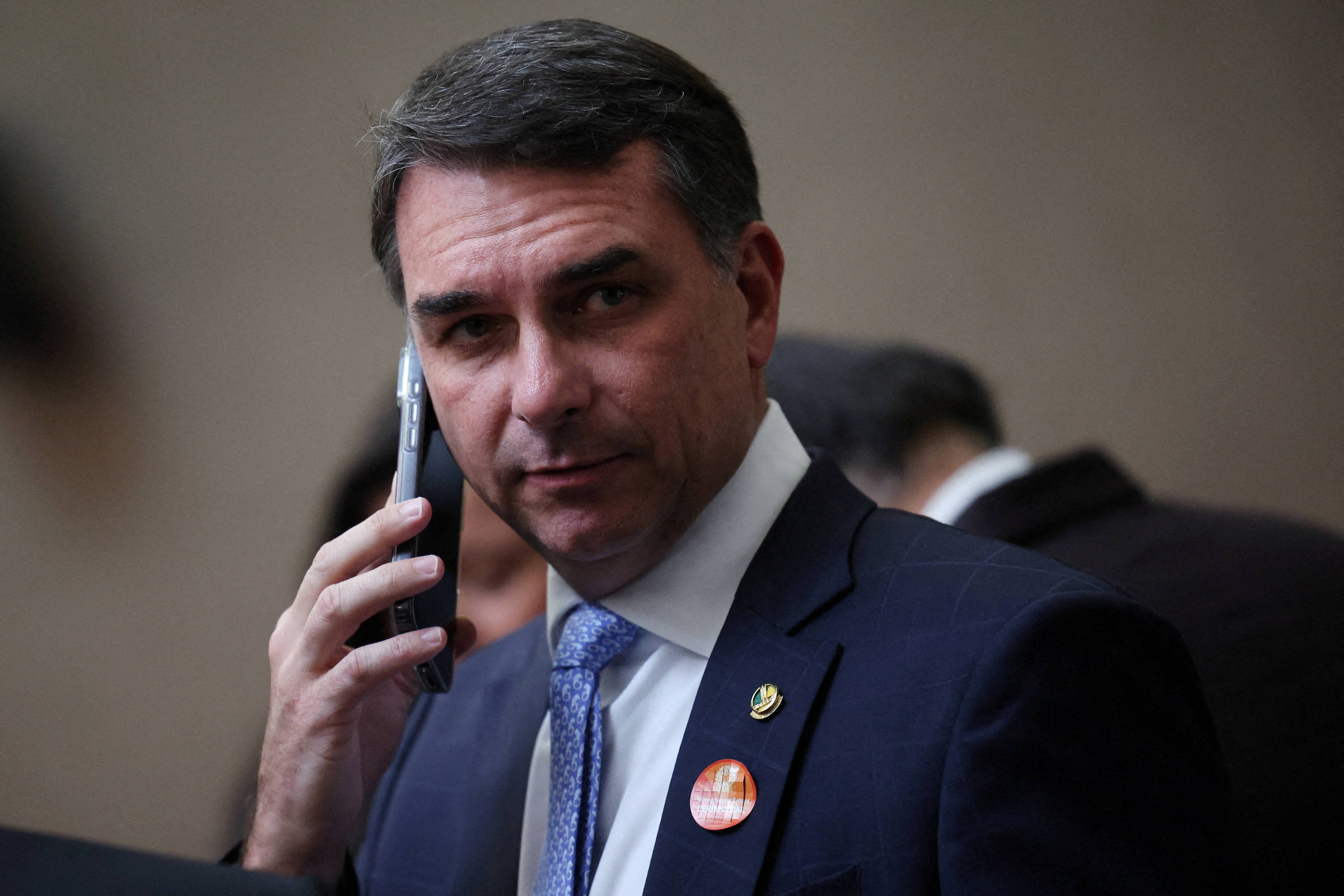 Homem de meia-idade com cabelo curto e escuro veste terno azul, camisa branca e gravata azul clara. Ele segura um celular próximo à orelha direita, olhando diretamente para a câmera. O fundo é desfocado, com outras pessoas parcialmente visíveis.