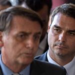 Dois homens em traje formal, o primeiro em primeiro plano desfocado e o segundo ao fundo focado, ambos com expressão séria. Ambiente interno com outras pessoas ao fundo.