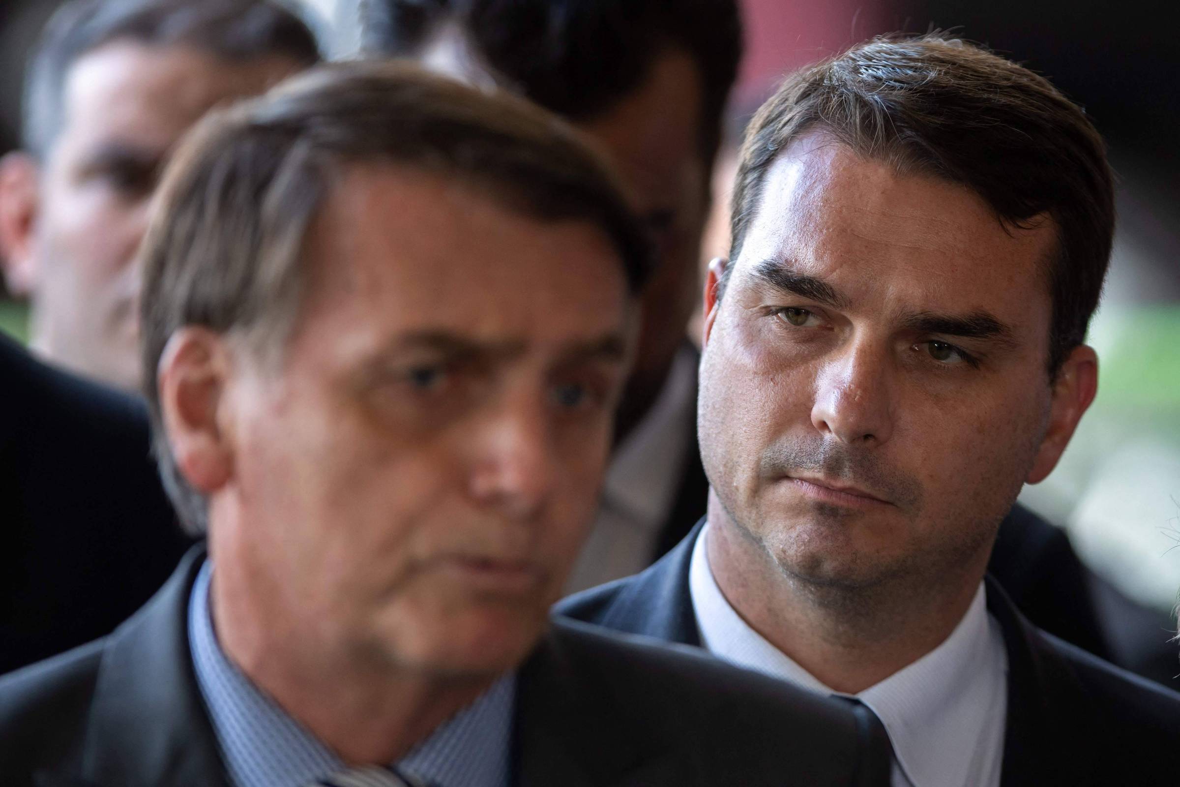 Dois homens em traje formal, o primeiro em primeiro plano desfocado e o segundo ao fundo focado, ambos com expressão séria. Ambiente interno com outras pessoas ao fundo.