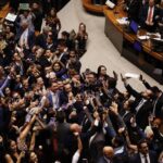 Grande grupo de deputados reunidos no plenário de uma casa legislativa, muitos tirando fotos com celulares e comemorando. Alguns parlamentares estão sentados nas mesas ao fundo, observando a movimentação.