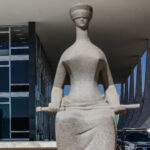 A imagem mostra uma escultura da Justiça, representada por uma figura feminina com os olhos vendados, segurando uma balança. A estátua está em primeiro plano, enquanto ao fundo se vê um edifício moderno com janelas de vidro e colunas. Há também um carro estacionado próximo à escultura e uma bandeira do Brasil visível.