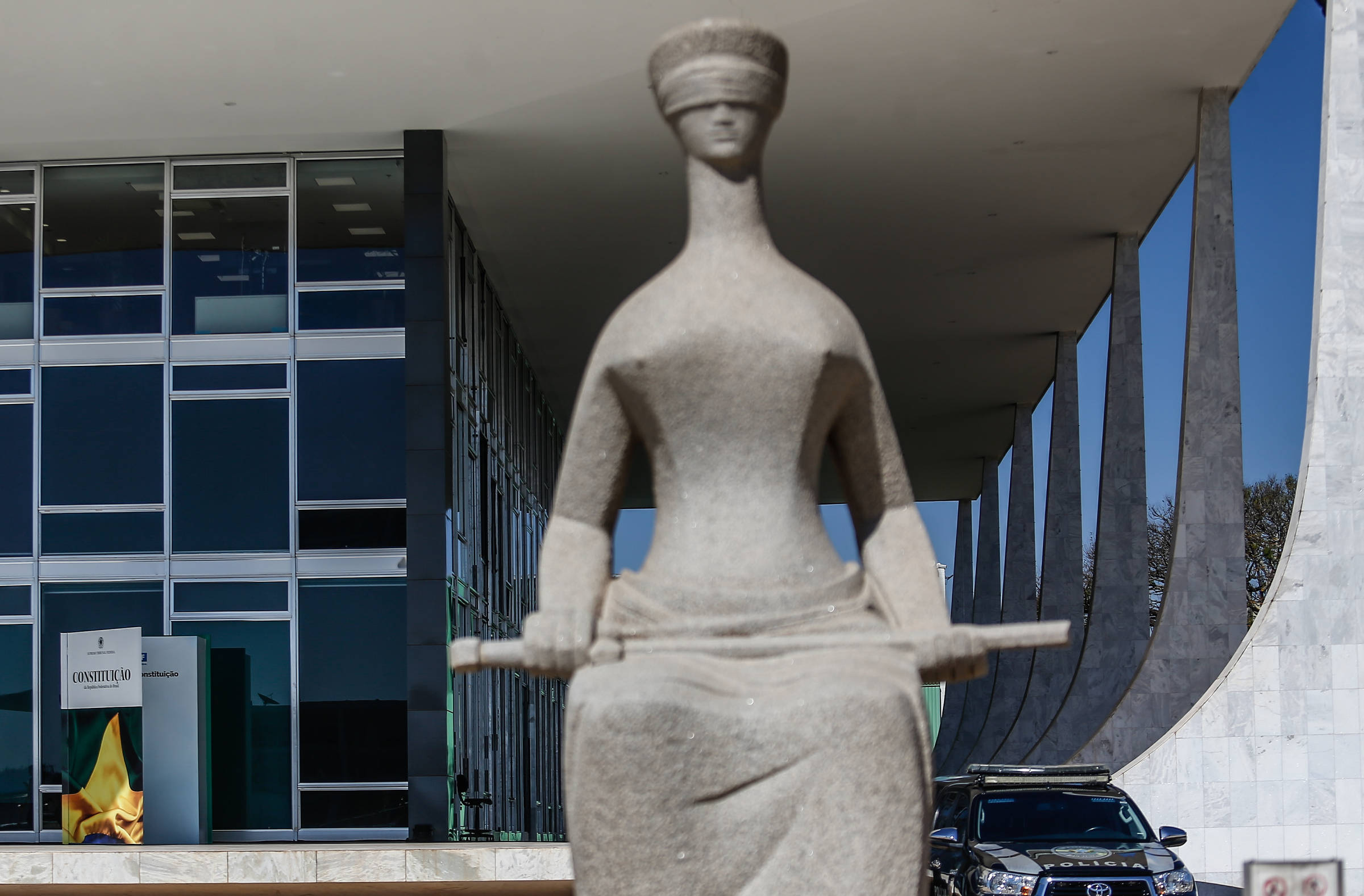 A imagem mostra uma escultura da Justiça, representada por uma figura feminina com os olhos vendados, segurando uma balança. A estátua está em primeiro plano, enquanto ao fundo se vê um edifício moderno com janelas de vidro e colunas. Há também um carro estacionado próximo à escultura e uma bandeira do Brasil visível.