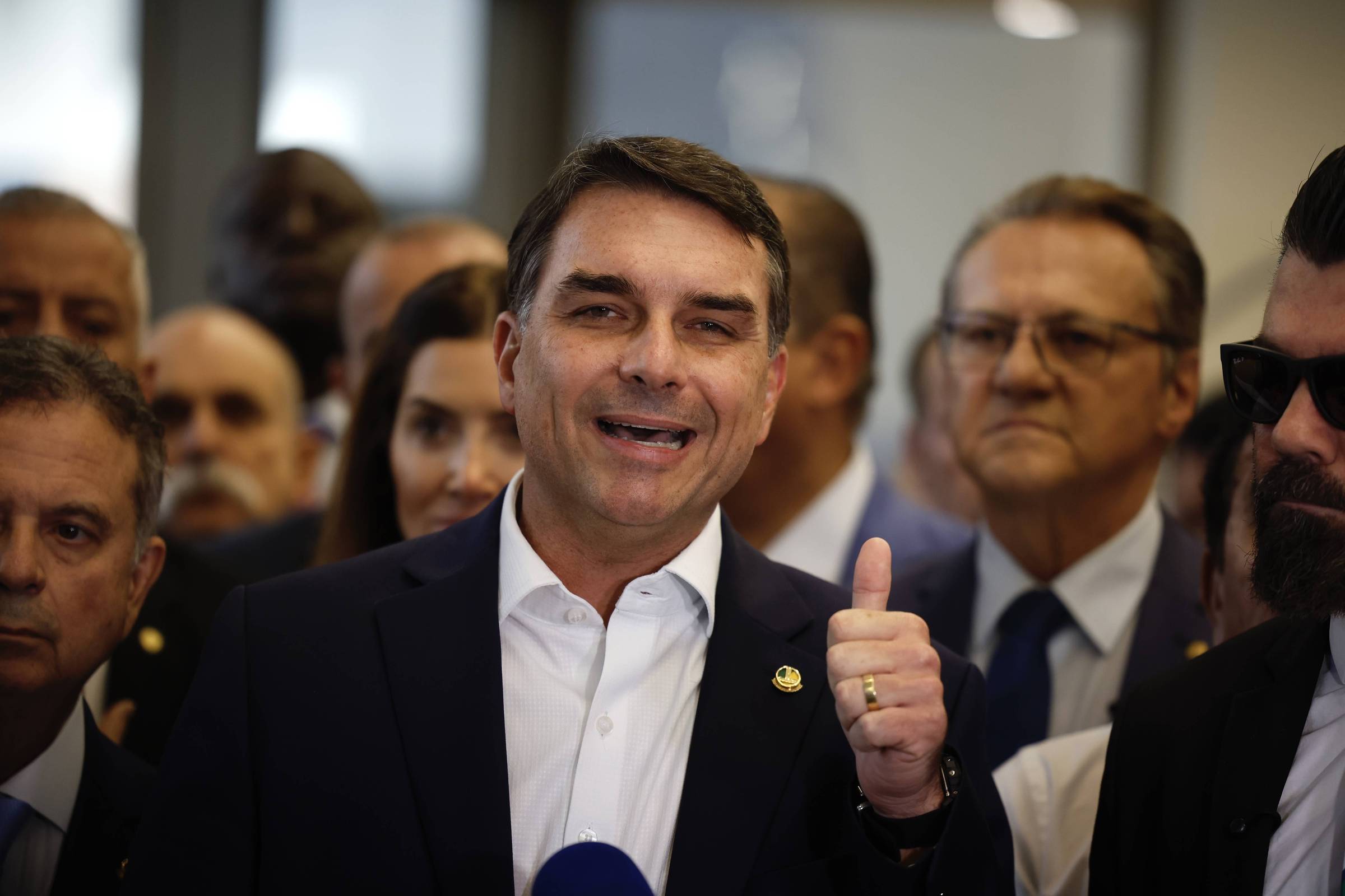 Homem de terno escuro e camisa branca sorri e levanta o polegar direito em gesto de aprovação, cercado por várias pessoas em ambiente interno.