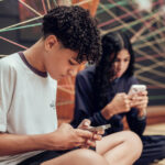 Dois jovens sentados no chão, cada um olhando para seu celular. Ao fundo, fios coloridos entrelaçados formam um padrão geométrico. O jovem à frente veste camiseta branca e o outro, ao fundo, veste roupa escura.