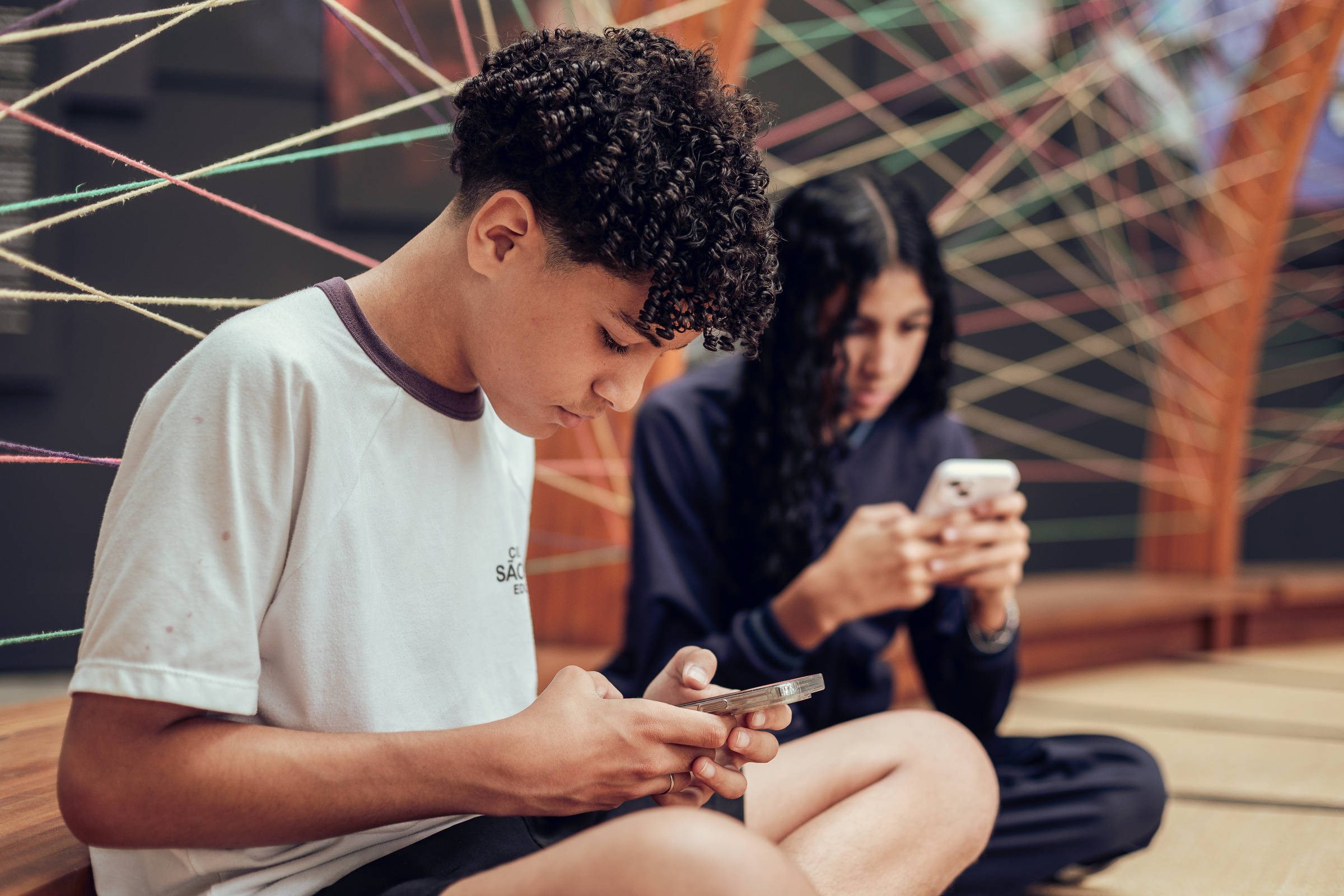 Dois jovens sentados no chão, cada um olhando para seu celular. Ao fundo, fios coloridos entrelaçados formam um padrão geométrico. O jovem à frente veste camiseta branca e o outro, ao fundo, veste roupa escura.