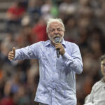Homem de cabelos grisalhos e barba, vestindo camisa azul clara, fala ao microfone em evento com público. Duas pessoas sentadas em cadeiras brancas ao seu lado, uma delas com colete azul claro e cabelos cacheados. Plateia visível ao fundo, ambiente interno.