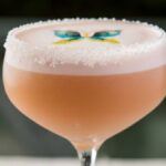 Taça de coquetel com bebida rosa clara e espuma branca na superfície. Decoração colorida em forma de borboleta sobre a espuma. Fundo desfocado com tons verdes.