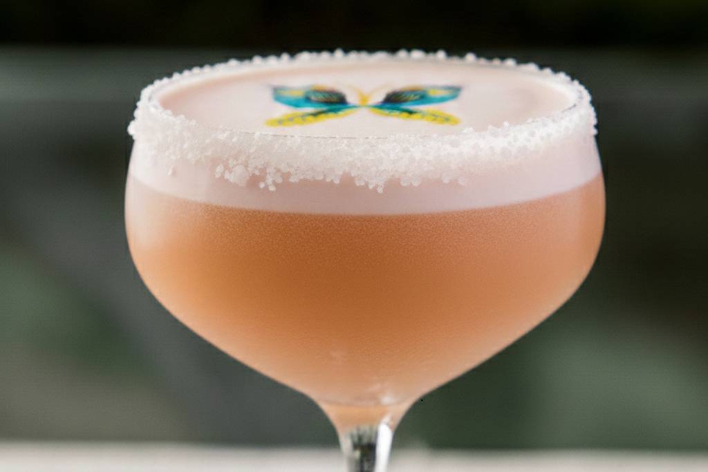 Taça de coquetel com bebida rosa clara e espuma branca na superfície. Decoração colorida em forma de borboleta sobre a espuma. Fundo desfocado com tons verdes.